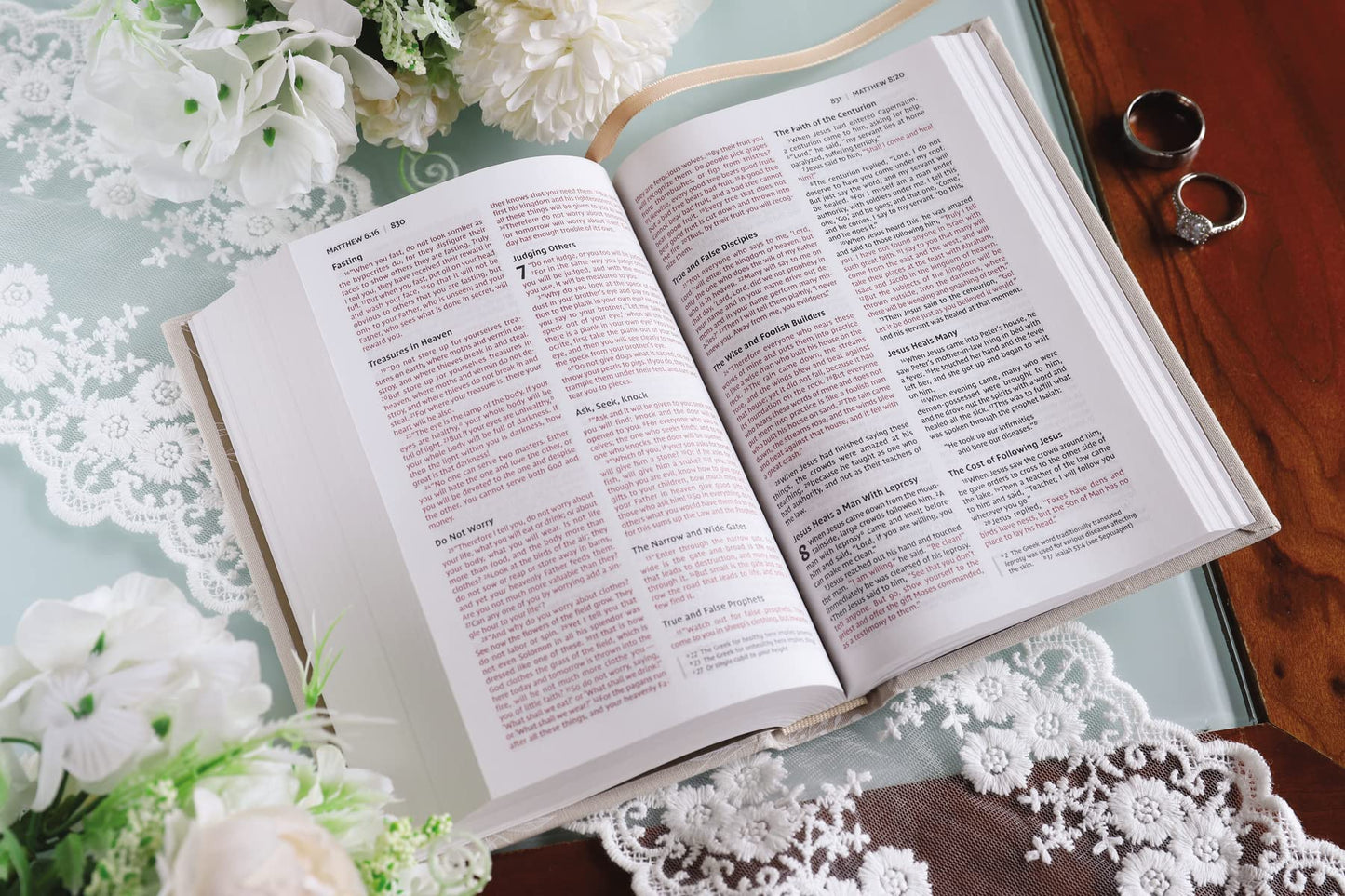 Una Biblia para novias de la NIV, abierta, con texto en rojo y negro, descansa sobre una mesa cubierta de encaje junto a dos anillos, flores blancas y una superficie de madera: una escena elegante, perfecta como regalo de boda o para una fiesta de despedida de soltera.