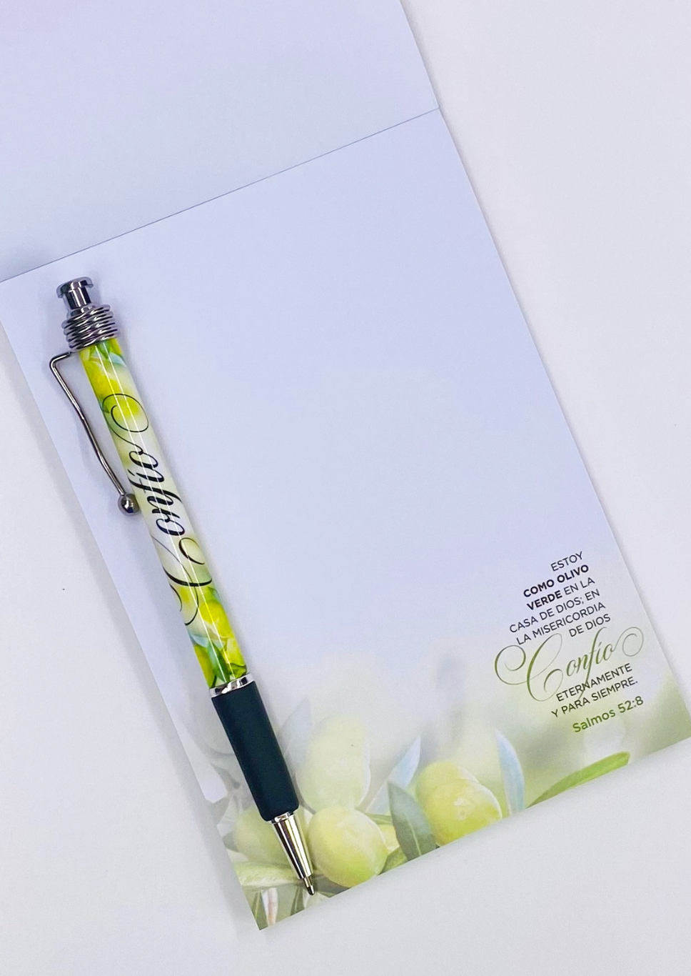 Un bolígrafo descansa sobre un cuaderno cristiano abierto, con un diseño inspirado en el olivo y un versículo bíblico del Salmo 52:8 en español impreso en la esquina inferior derecha.