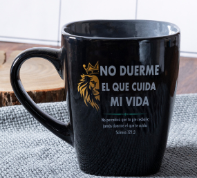 Taza de cerámica negra con un dibujo de un león dorado y un texto en español que reza: «No duerme el que cuida mi vida», junto con la referencia a un versículo bíblico, lo que le aporta un toque de fe y un estilo único, expuesta sobre un paspartú de tela.