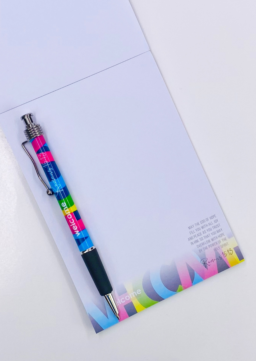 Un bolígrafo de colores descansa sobre un cuaderno abierto con versículos bíblicos, en el que aparece la palabra «bienvenido» impresa en ambos artículos. El cuaderno incluye un breve poema impreso y Romanos 15:13 en la esquina inferior derecha, lo que lo convierte en un regalo cristiano muy especial.