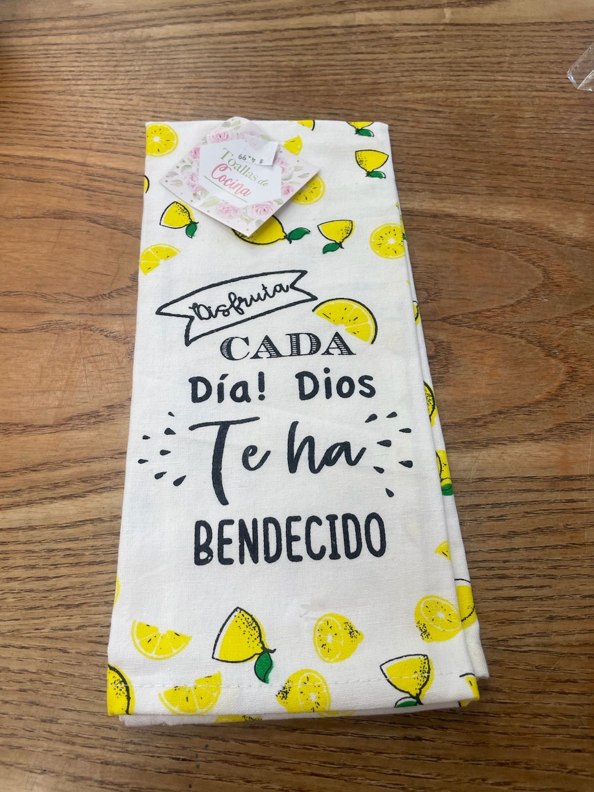 Sobre una superficie de madera hay un paño de cocina doblado con alegres ilustraciones de limones y un texto en español que dice: «¡Disfruta cada día! Dios te ha bendecido».