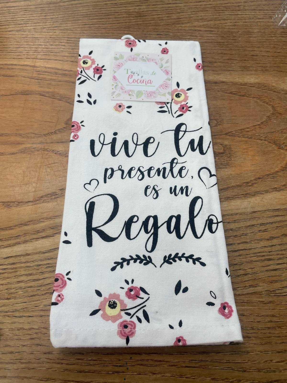 Un paño blanco con motivos florales rosas y la frase en español «Vive tu presente, es un regalo», colocado con elegancia sobre una superficie de madera.