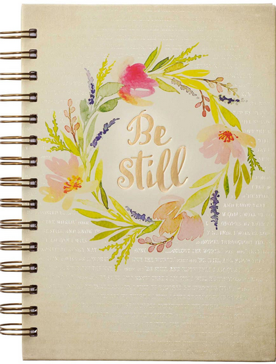 Un cuaderno con espiral, con un diseño de corona de flores y la frase «Be still» en letras doradas en la portada, con páginas rayadas para tomar notas fácilmente.