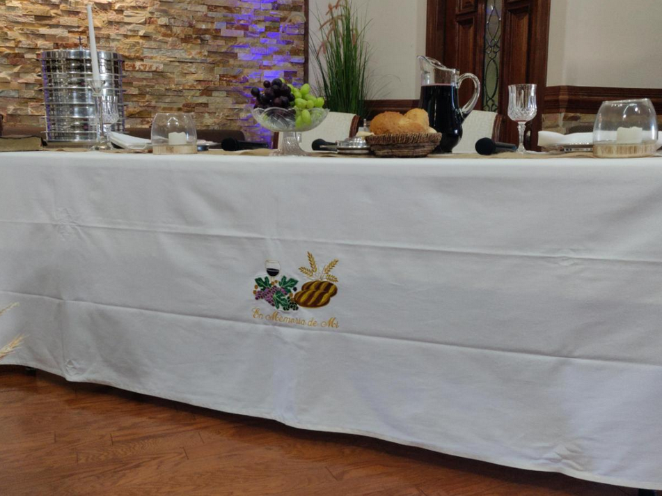 Una mesa con un mantel blanco para la mesa de la Cena del Señor, en la que se aprecian pan, uvas, una jarra, vasos, bandejas apiladas y una cesta, situada en una estancia con suelo de madera y detalles de piedra en las paredes.