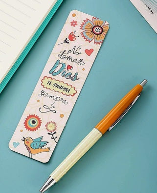 Un colorido marcapáginas con flores, un pájaro y el reconfortante texto en español «No temas, Dios te ayudará siempre» descansa junto a un bolígrafo naranja y blanco sobre un escritorio azul, ofreciendo un suave recordatorio de ayuda y esperanza.