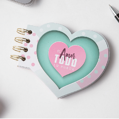 Un cuaderno de tapa dura con forma de corazón y troquelado, con un corazón rosa en la portada, en el que figura el texto bíblico en español: «El amor todo lo puede».