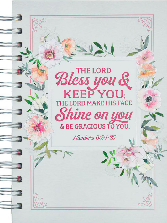 Un cuaderno con espiral, decorado con motivos florales y un versículo bíblico: «Que el Señor te bendiga y te proteja... Números 6:24-25», impreso en rosa y rojo en la portada, con páginas rayadas que contienen pasajes de las Escrituras en su interior.