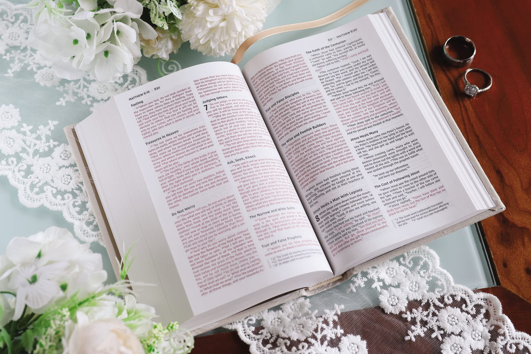 Una Biblia para novias de la NIV, abierta, con texto en rojo y negro, descansa sobre una mesa cubierta de encaje junto a dos anillos, flores blancas y una superficie de madera: una escena elegante, perfecta como regalo de boda o para una fiesta de despedida de soltera.