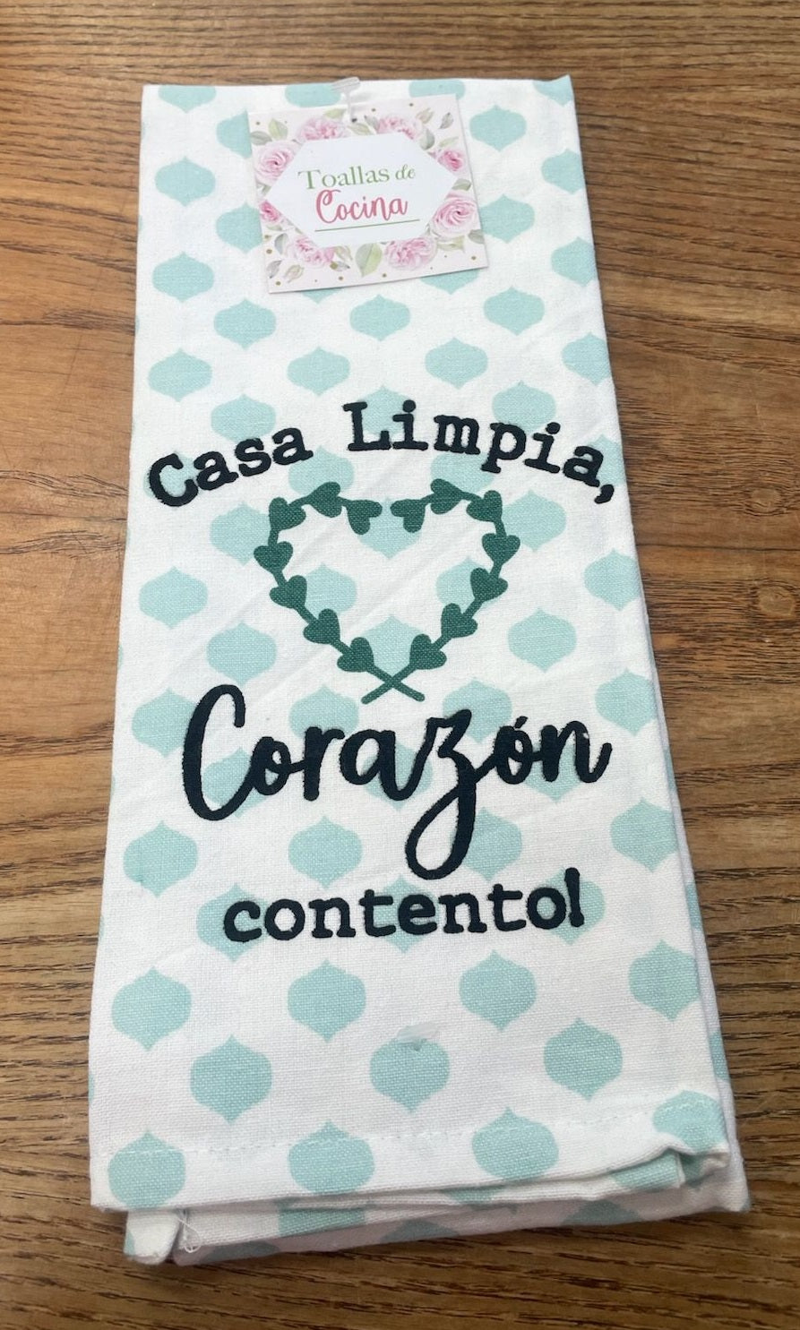 Un paño de cocina doblado con un estampado azul claro y un texto en español: «¡Casa limpia, corazón contento!», con un diseño de un corazón en el centro, perfecto para añadir alegría a tu rutina de limpieza del hogar, colocado sobre una superficie de madera.