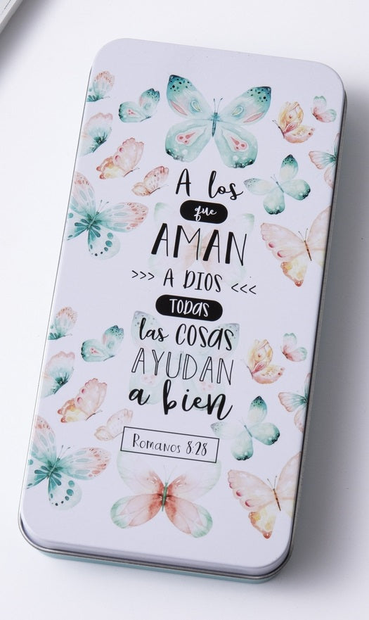 Una caja metálica rectangular decorada con coloridas ilustraciones de mariposas y un texto en español tomado de Romanos 8:28: «A los que aman a Dios, todas las cosas les ayudan a bien».