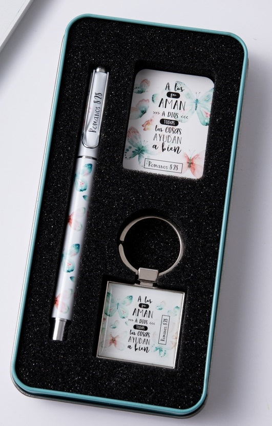 Set de regalo compuesto por un bolígrafo, un llavero y una tarjeta, todos ellos decorados con motivos florales y un texto en español que incluye un versículo bíblico de Romanos 8:28, presentados de forma elegante en una caja metálica forrada de espuma.