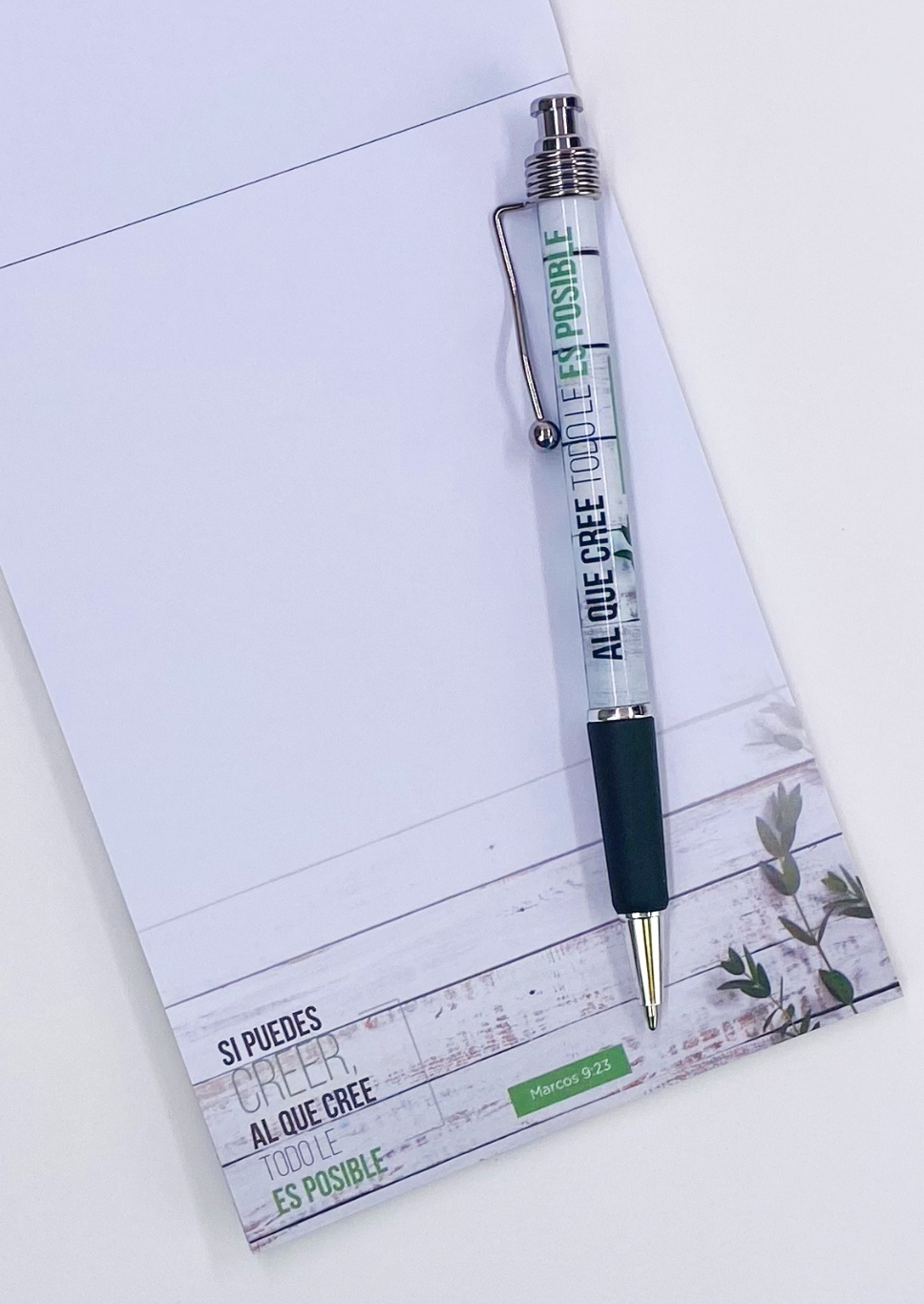 Un bolígrafo transparente con texto en español descansa sobre un cuaderno cristiano decorado con un motivo vegetal. El texto dice: «SI PUEDES CREER, AL QUE CREE TODO LE ES POSIBLE» —versículo bíblico, Marcos 9:23.