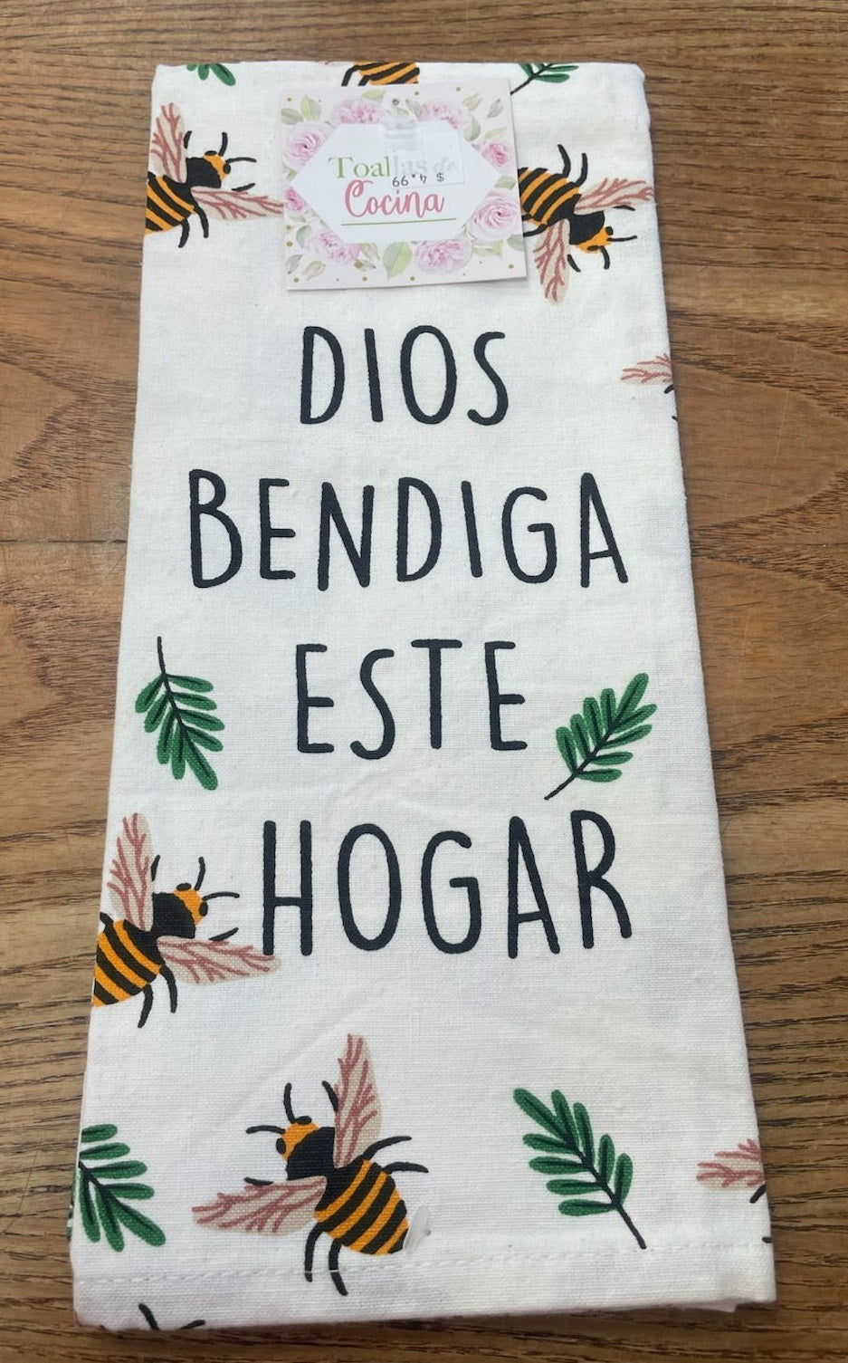 Un paño de cocina blanco con la frase «Dios bendiga este hogar» en español, decorado con abejas y hojas verdes, colocado sobre una superficie de madera.