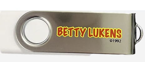 Una memoria USB plateada con el texto «BETTY LUKENS ©1992» impreso en letras amarillas y rojas en la parte metálica, ideal para guardar actividades bíblicas o láminas para colorear.