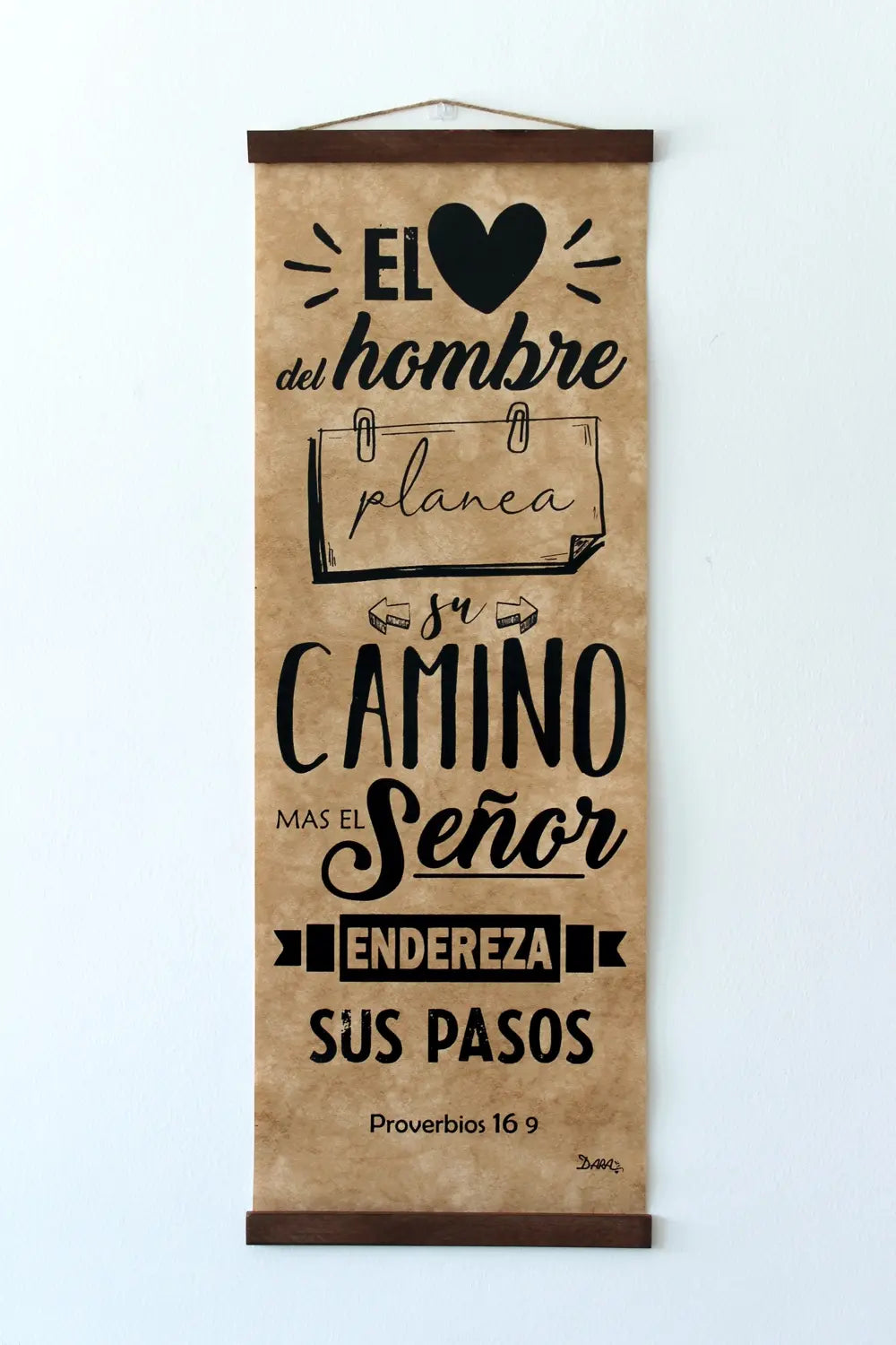 Un rollo vertical para colgar en la pared muestra el versículo de la Biblia en español de Proverbios 16:9 en letra negra sobre papel marrón, con tipografías decorativas e ilustraciones pequeñas, y constituye una encantadora decoración cristiana similar a un cuadro de madera.