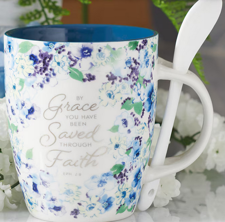 Una taza de cerámica con un diseño floral azul y una cucharita blanca en el asa, en la que se lee, en elegante letra cursiva: «Por gracia habéis sido salvados mediante la fe» (Ef 2,8).