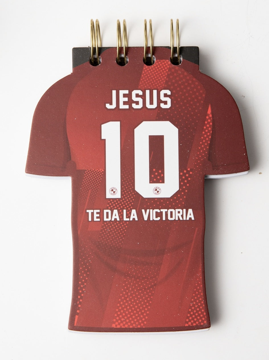 Una libreta troquelada con forma de camiseta de fútbol roja, con tapa dura y la frase «JESUS 10 TE DA LA VICTORIA» impresa en la portada, ideal para quienes buscan inspiración y pasajes bíblicos en su día a día.