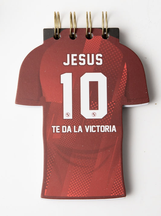 Una libreta troquelada con forma de camiseta de fútbol roja, con tapa dura y la frase «JESUS 10 TE DA LA VICTORIA» impresa en la portada, ideal para quienes buscan inspiración y pasajes bíblicos en su día a día.