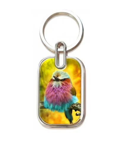 Un llavero con la imagen de un pájaro de colores, con plumas moradas, azules y turquesas, posado en una rama sobre un fondo amarillo, inspirado en 1 Juan 4, 19: «Amamos a Dios porque Él nos amó primero».