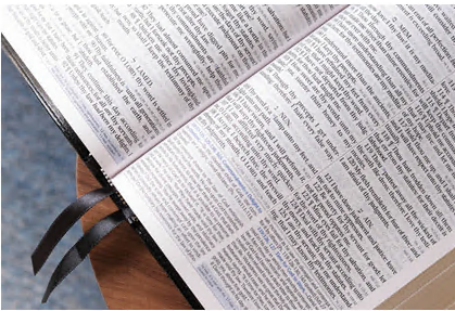 Una Biblia de letra grande, abierta y con cintas negras, yace sobre una superficie de madera, mostrando el texto impreso de la Versión del Rey Jacobo en ambas páginas visibles.