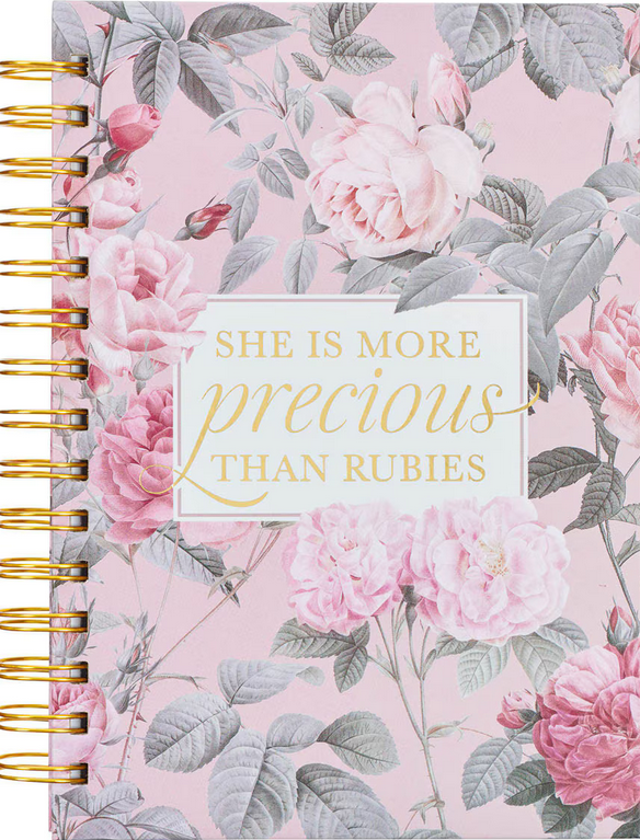 Un cuaderno rosa con motivos florales, encuadernado con espiral, con páginas rayadas y la frase «Ella es más preciosa que los rubíes» impresa en dorado en la tapa dura.