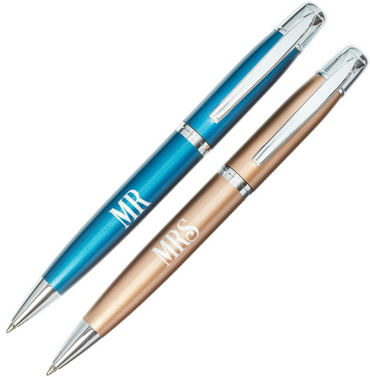 Dos bolígrafos metálicos de mecanismo giratorio, uno azul con la etiqueta «MR» y otro dorado con la etiqueta «MRS», ambos con clips y detalles plateados, se presentan colocados en paralelo dentro de una elegante caja de regalo.