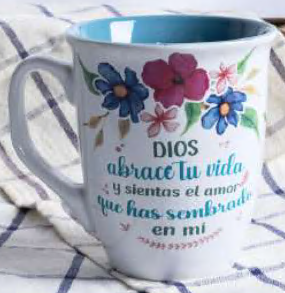 Una taza blanca de 14 oz con motivos florales y un texto en español que dice: «Dios abrace tu vida y sientas el amor que has sembrado en mí», descansa sobre un mantel a cuadros. Esta taza, con el interior de color, aporta un toque de elegancia, lo que la hace perfecta para tu café o té diario.