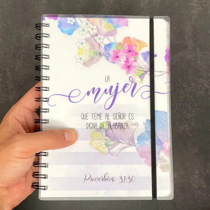 Una mano sostiene una agenda cristiana con motivos florales y texto en español: «La mujer que teme al Señor es digna de alabanza». Proverbios 31:30. Este cuaderno con encuadernación en espiral es perfecto para tener siempre a mano un versículo diario.