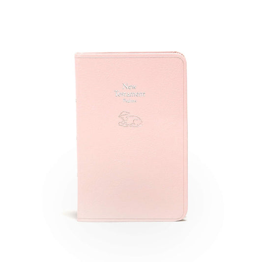 Un ejemplar del Nuevo Testamento para bebés de la versión KJV, de color rosa claro, que incluye los Salmos y una ilustración de un cordero en relieve plateado en la portada, representado de pie sobre un fondo blanco; ideal como regalo para la dedicatoria de un bebé o como Biblia de recuerdo.