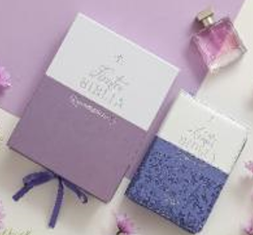 Dos Biblias «Reina Valera 1960», de color morado y blanco, en formato portátil, se exhiben junto a un frasco de perfume rosa y unas pequeñas flores moradas sobre un fondo lavanda: perfectas para una Biblia de quinceañera.