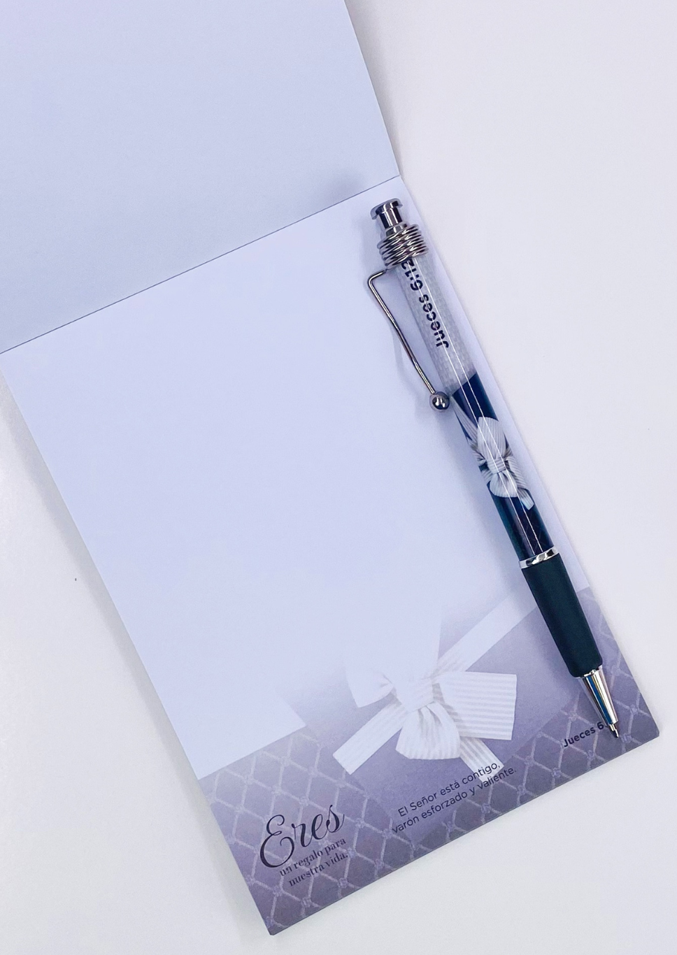 Un cuaderno cristiano con un bolígrafo incluido encima. La parte inferior del cuaderno presenta un texto decorativo y un lazo blanco sobre un fondo con un estampado gris claro.