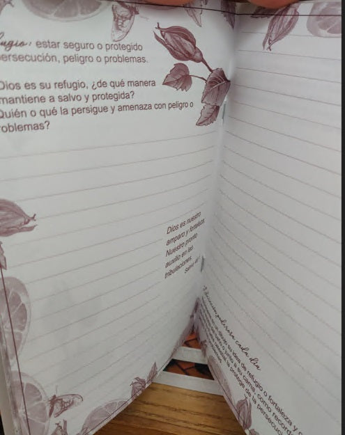 Un cuaderno de oración abierto, con adornos florales, muestra páginas rayadas y un texto en español sobre el refugio, la protección y preguntas para la reflexión.