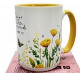 Taza blanca con el interior y el asa amarillos, decorada con ilustraciones de flores amarillas, hojas verdes, una mariposa y un texto en el lateral.
