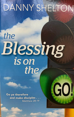 La portada del libro titulado *The Blessing is on the GO!*, de Danny Shelton, muestra un semáforo en verde sobre un cielo azul junto a un versículo bíblico, Mateo 28:19, que destaca un ministerio inspirado en los principios bíblicos.