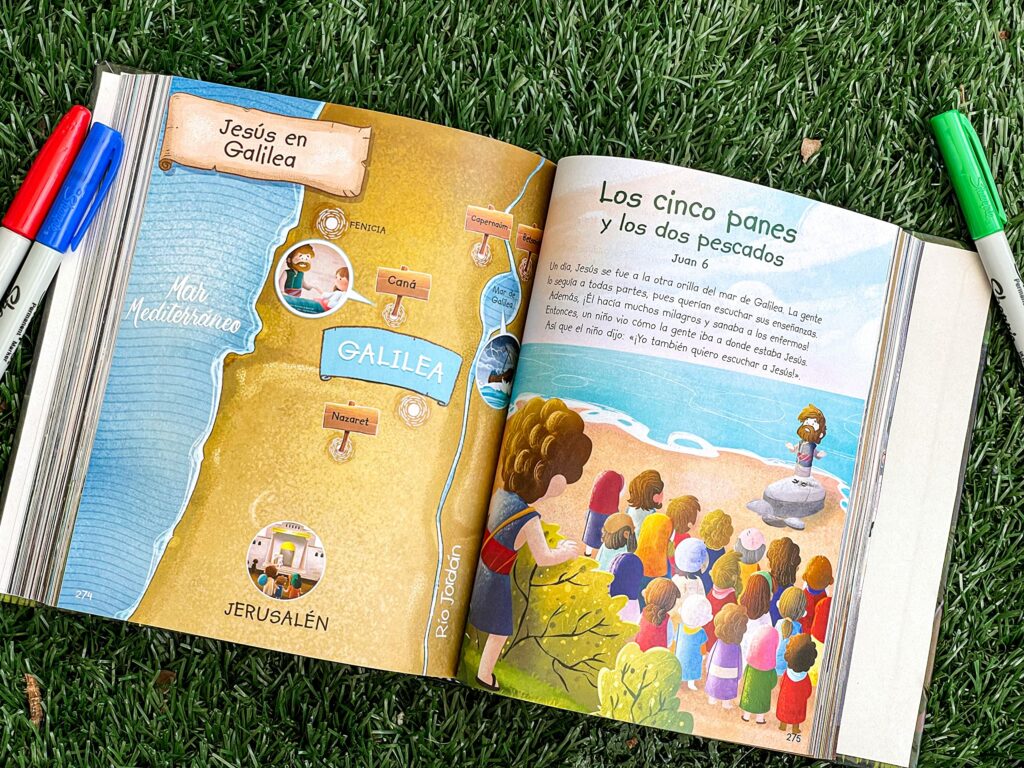 Un libro ilustrado abierto, destinado a niños de entre 6 y 8 años, descansa sobre la hierba; en él se ve un mapa de Galilea y una historia bíblica ilustrada en la que Jesús alimenta a una multitud, con rotuladores cerca.