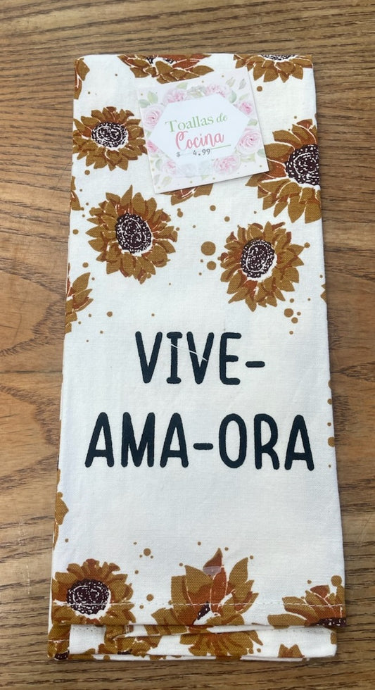 Un paño de cocina blanco con estampados de girasoles y las palabras motivadoras «Ora, Vive, Ama» en negro, colocado sobre una superficie de madera. En la parte superior lleva una etiqueta en la que se lee «Toallas de Cocina».