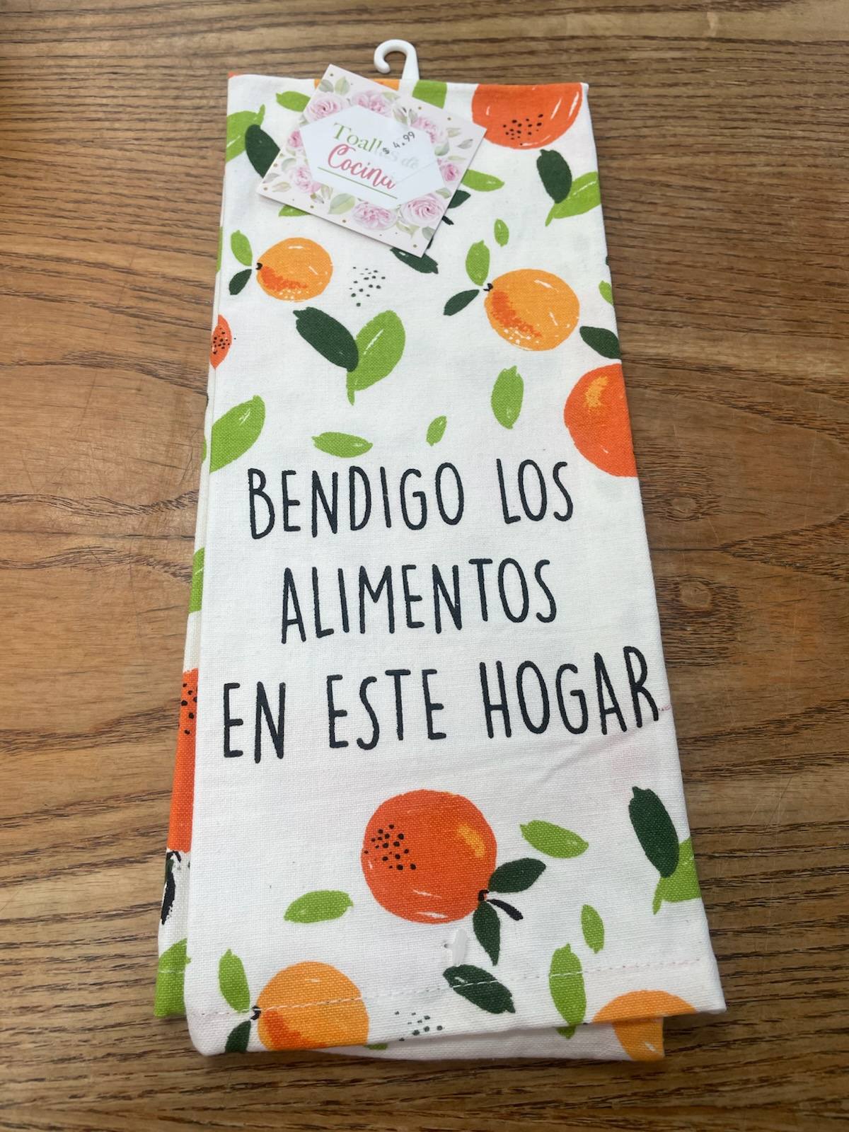 Sobre una superficie de madera descansa un paño de cocina blanco doblado, con dibujos de frutas naranjas y verdes y la frase en español «Bendigo los alimentos en este hogar».