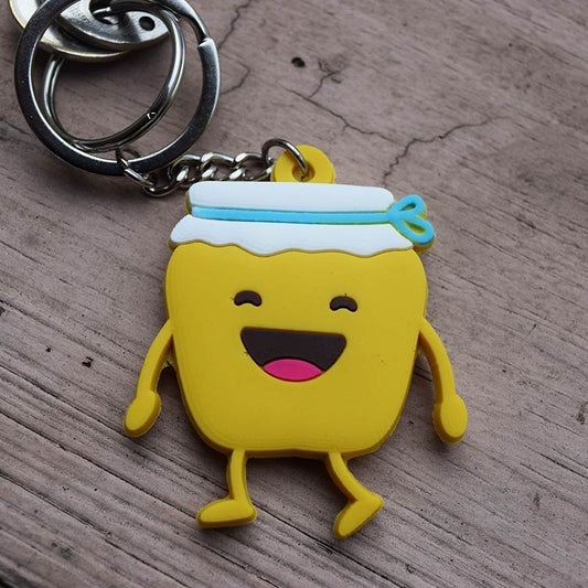 Divertido llavero con un frasco amarillo de estilo caricaturesco, una carita sonriente, una tapa blanca y una cinta azul, acompañado de la cita del Salmo 119:103; un regalo cristiano ideal sobre una base de madera.