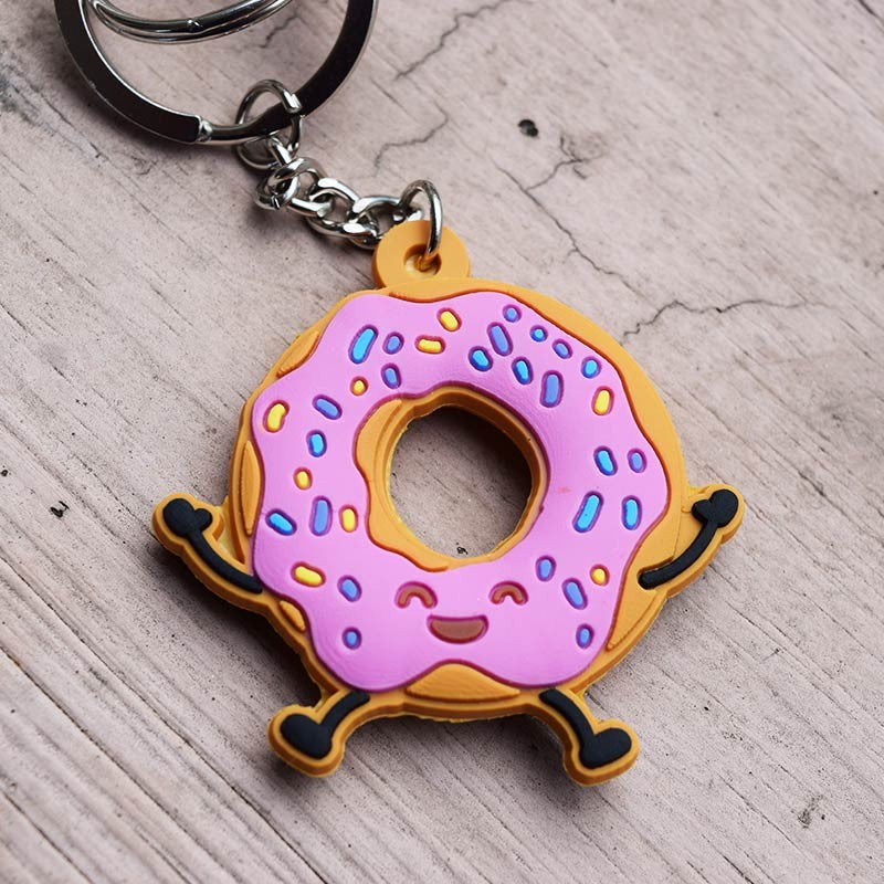 Un llavero divertido con una rosquilla de dibujos animados con glaseado rosa, virutas de colores y una cara sonriente, apoyada sobre una superficie de madera; perfecto como regalo cristiano para cualquier ocasión.