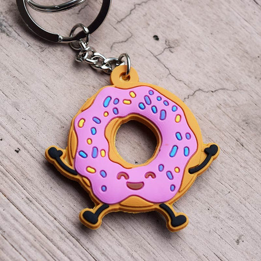 Un llavero divertido con una rosquilla de dibujos animados con glaseado rosa, virutas de colores y una cara sonriente, apoyada sobre una superficie de madera; perfecto como regalo cristiano para cualquier ocasión.