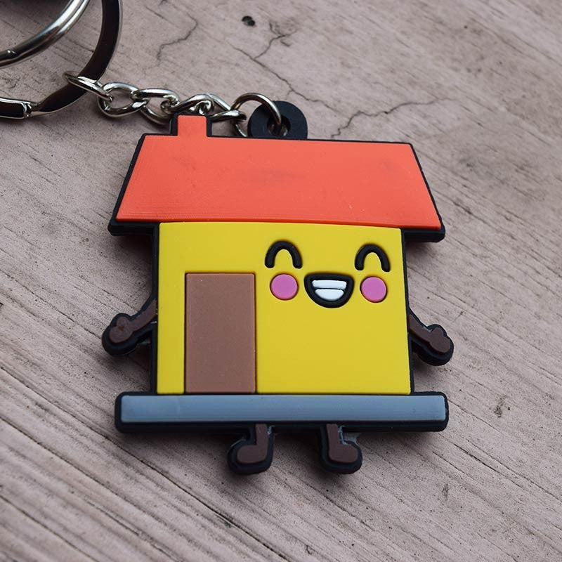 Un llavero divertido con la forma de una casita de dibujos animados con tejado rojo, paredes amarillas, una cara sonriente y unas patitas, colocado sobre una superficie de madera. Perfecto como regalo cristiano inspirado en Josué 24:15.