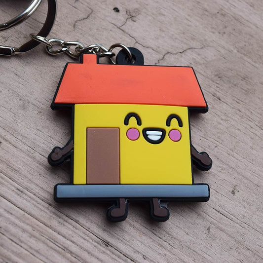 Un llavero divertido con la forma de una casita de dibujos animados con tejado rojo, paredes amarillas, una cara sonriente y unas patitas, colocado sobre una superficie de madera. Perfecto como regalo cristiano inspirado en Josué 24:15.