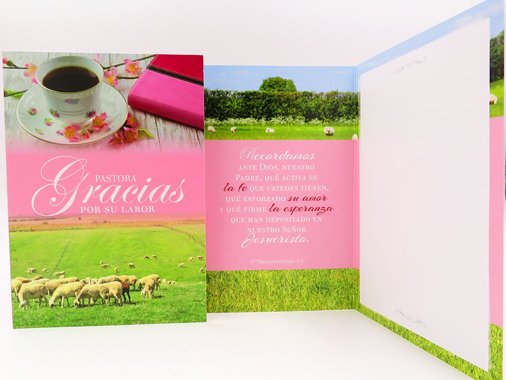 Una tarjeta de alta calidad con una taza de té, un platillo y un cuaderno con motivos florales en la portada, además de ovejas en un prado verde bajo un cielo azul; el texto en español es de color rosa en el interior. Incluye un sobre y una bolsa protectora para guardarla en buen estado.