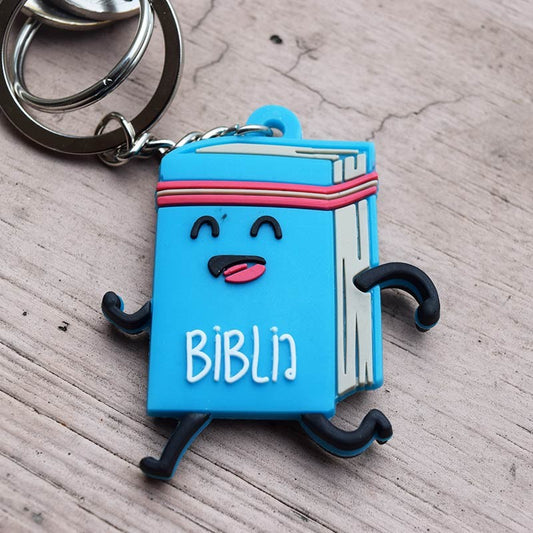 Un llavero divertido que representa un libro azul animado, con brazos y piernas sonrientes, con la etiqueta «BIBLIN», apoyado sobre una superficie de madera; un regalo cristiano ideal para llevar contigo el versículo de Hebreos 12:1.