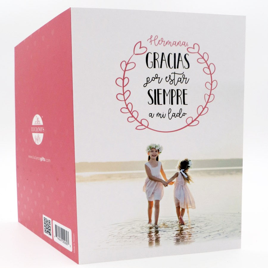 Una caja de regalo rosa y blanca muestra a dos niñas con vestidos blancos cogidas de la mano en la playa, con un texto en español que dice: «Hermana, gracias por estar siempre a mi lado». Incluye una tarjeta de alta calidad y una bolsa protectora.
