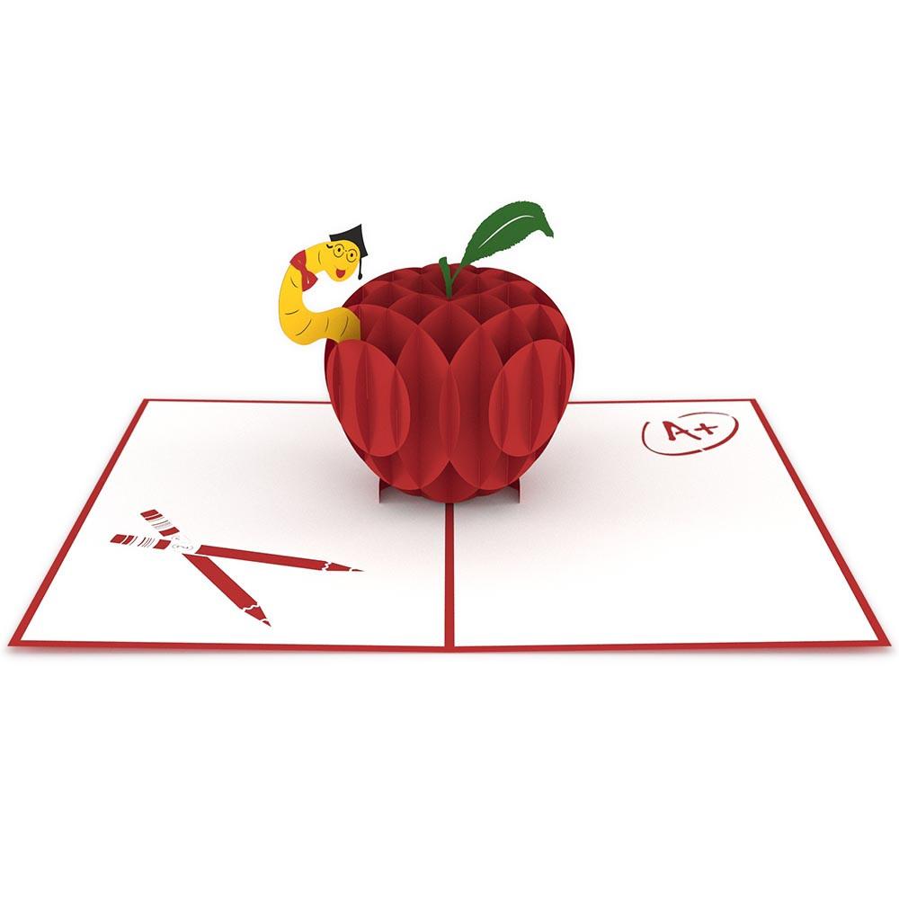 Una tarjeta pop-up en 3D de alta calidad muestra una manzana roja con un gusano, colocada sobre una tarjeta abierta en la que se ven dos lápices y una nota de A+.