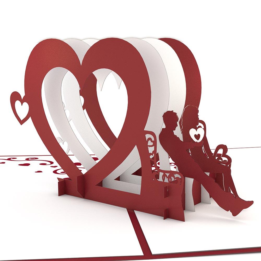 Escultura de papel en tonos rojos y blancos que representa a dos personas sentadas en un banco, enmarcada por corazones entrelazados con pequeños recortes en forma de corazón, colocada sobre una superficie plana. Perfecta como tarjeta de 5 x 7 o tarjeta de alta calidad para ocasiones especiales.
