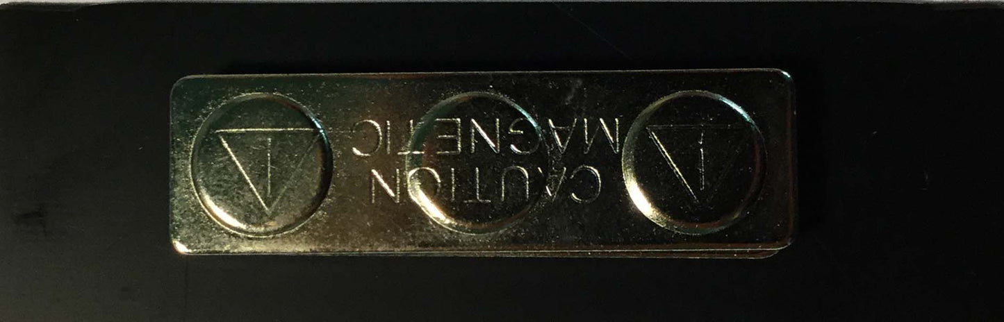 A una superficie negra se ha fijado una placa metálica distintiva con cierre magnético en la que figura el texto «CAUTION MAGNETIC» y unos símbolos triangulares.