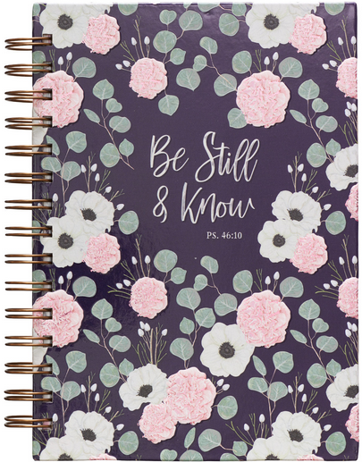 Un cuaderno floral con encuadernación de alambre en el que se lee «Be Still & Know PS. 46:10» en una cubierta de color morado oscuro, decorado con flores rosas y blancas y hojas verdes, con páginas rayadas para tus pensamientos y reflexiones.