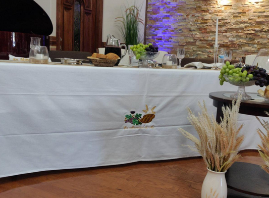 Un mantel blanco para la mesa de la Cena del Señor, con uvas y pan bordados, cubre una mesa en la que hay pan, uvas, agua, una jarra de café y velas; al fondo se ven plantas decorativas y una pared de piedra.